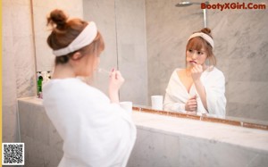 A woman in a white robe brushing her teeth in front of a mirror.