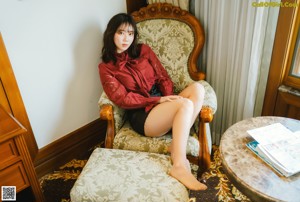 A woman sitting on a bed wearing a red shirt and black shorts.